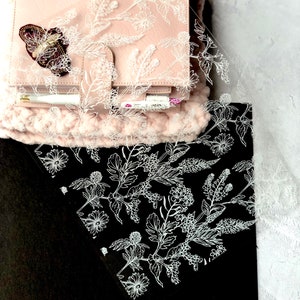 May include: A clear acrylic planner sheet with a white floral design. The sheet is placed on top of a black planner and a pink planner with a butterfly embellishment.