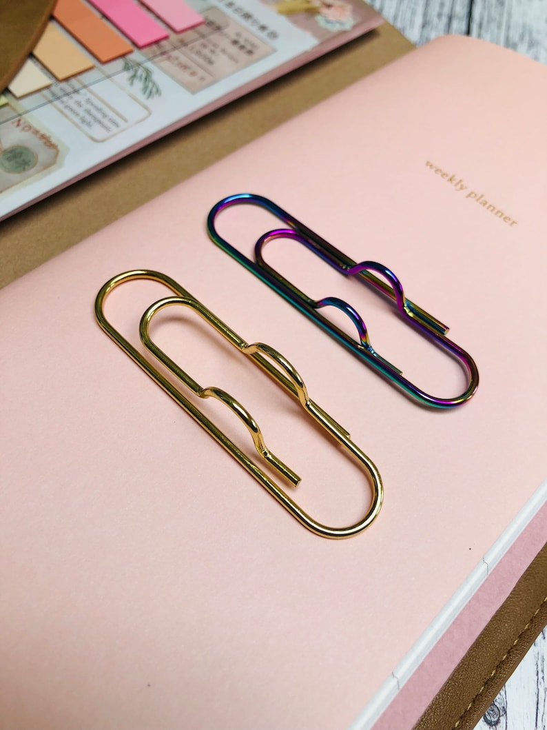 May include: Two paper clips, one gold and one iridescent, on a pink surface with the text "weekly planner" printed in white.