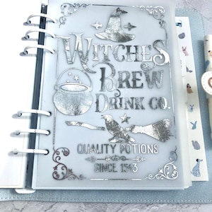 May include: A silver foiled planner cover with a witch's hat, cauldron, broom, and raven. The text reads "Witches Brew Drink Co. Quality Potions Since 1543."