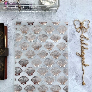 May include: A clear plastic sheet with a repeating pattern of gold foil seashells on a white marble background. The sheet is designed for use in a planner or journal.