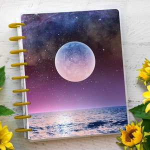 Puede incluir: Un cuaderno de espiral cerrado con una cubierta colorida que presenta una gran luna llena sobre un océano azul oscuro con un cielo estrellado. La cubierta tiene un degradado de rosa, morado y azul.