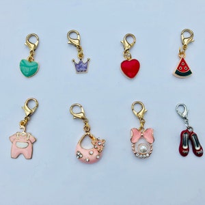 Puede incluir: Conjunto de diez colgantes con cierres de tono dorado. Los colgantes incluyen un corazón verde, una corona morada, un corazón rojo, una sandía, un mameluco rosa para bebé, un bolso rosa con pedrería, un bolso rosa con una perla y un lazo, y un par de zapatos rojos.