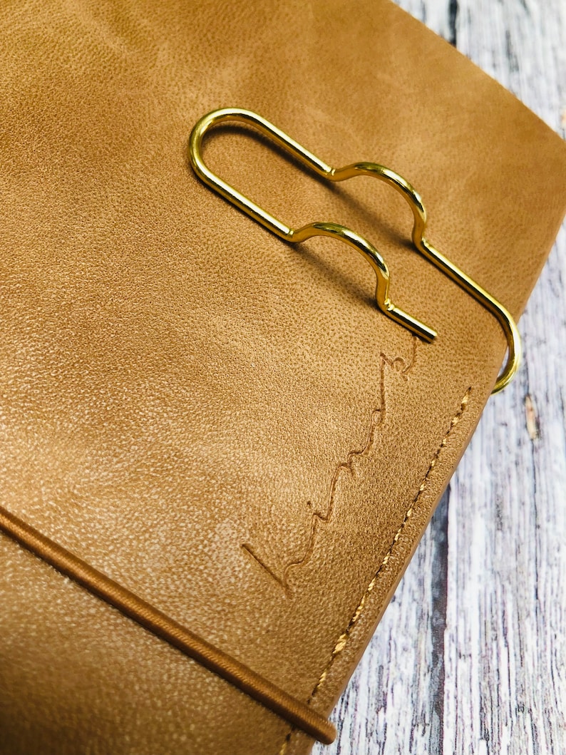 May include: A brown leather journal with a gold metal bookmark. The journal has a stitched edge and the text "Liminal" embossed on the cover.