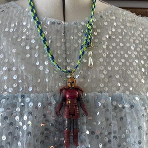 Op de afbeelding: Een handgemaakte ketting met een miniatuur actiefiguur hanger. De ketting heeft een blauw en neon groen gevlochten koord met een kleine parelbedel. De actiefiguur is rood en bruin met een gouden helm.
