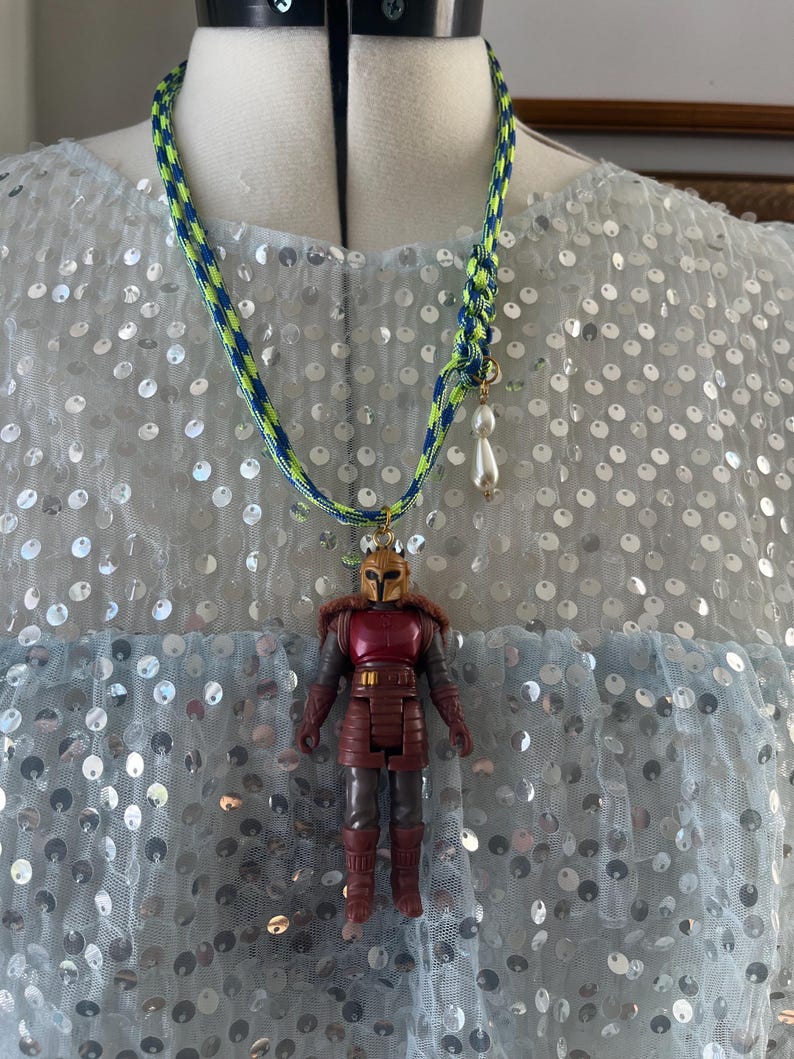 Op de afbeelding: Een handgemaakte ketting met een miniatuur actiefiguur hanger. De ketting heeft een blauw en neon groen gevlochten koord met een kleine parelbedel. De actiefiguur is rood en bruin met een gouden helm.