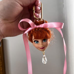 Puede incluir: Un llavero con cabeza de mu&ntilde;eca que presenta un personaje con cabello rojo, ojos azules y una cinta rosa. El llavero tiene un cierre dorado y un colgante de cristal transparente. La cabeza de la mu&ntilde;eca es el foco de este accesorio &uacute;nico.