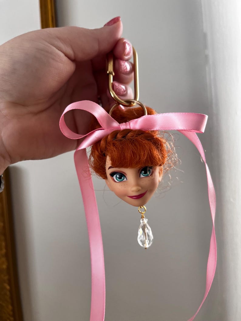 Puede incluir: Un llavero con cabeza de mu&ntilde;eca que presenta un personaje con cabello rojo, ojos azules y una cinta rosa. El llavero tiene un cierre dorado y un colgante de cristal transparente. La cabeza de la mu&ntilde;eca es el foco de este accesorio &uacute;nico.