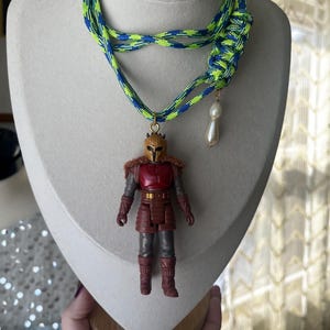 Op de afbeelding: Een unieke ketting met een miniatuur actiefiguur hanger. De ketting heeft een neon groen en blauw gevlochten koord. Een imitatie parel charme hangt naast het actiefiguur. Het actiefiguur draagt een rood en bruin outfit.