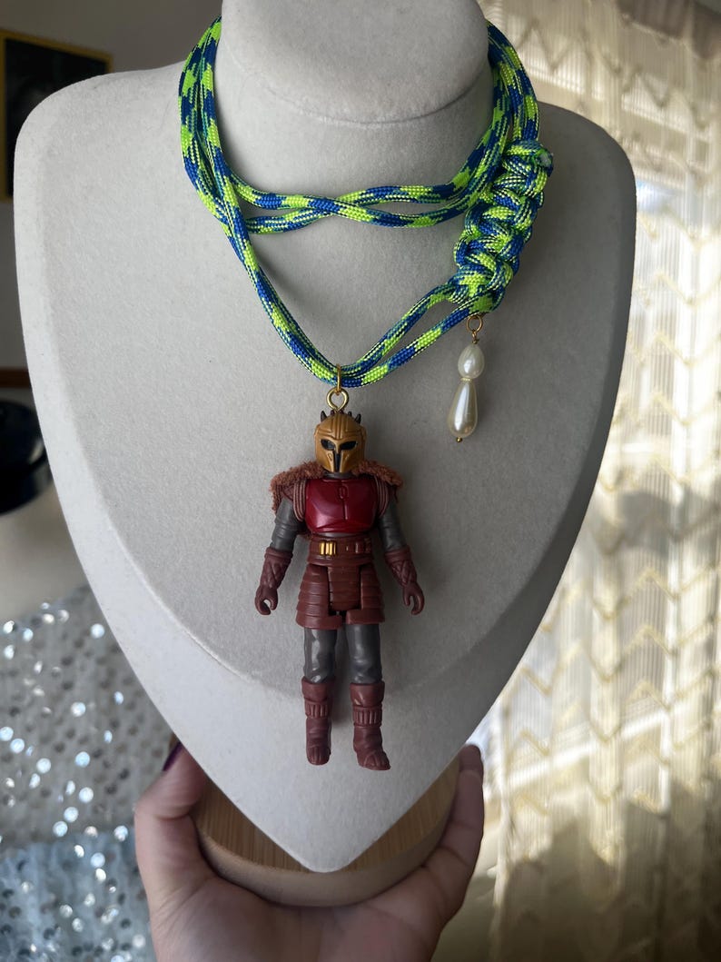 Op de afbeelding: Een unieke ketting met een miniatuur actiefiguur hanger. De ketting heeft een neon groen en blauw gevlochten koord. Een imitatie parel charme hangt naast het actiefiguur. Het actiefiguur draagt een rood en bruin outfit.