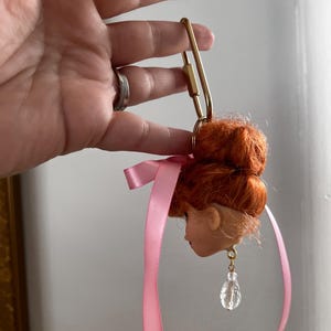 Puede incluir: Una cabeza de mu&ntilde;eca con pelo rojo y una cinta rosa, sujeta a un cierre dorado. La cabeza de la mu&ntilde;eca tiene un pendiente transparente en forma de l&aacute;grima. El cierre est&aacute; sujeto por una mano.