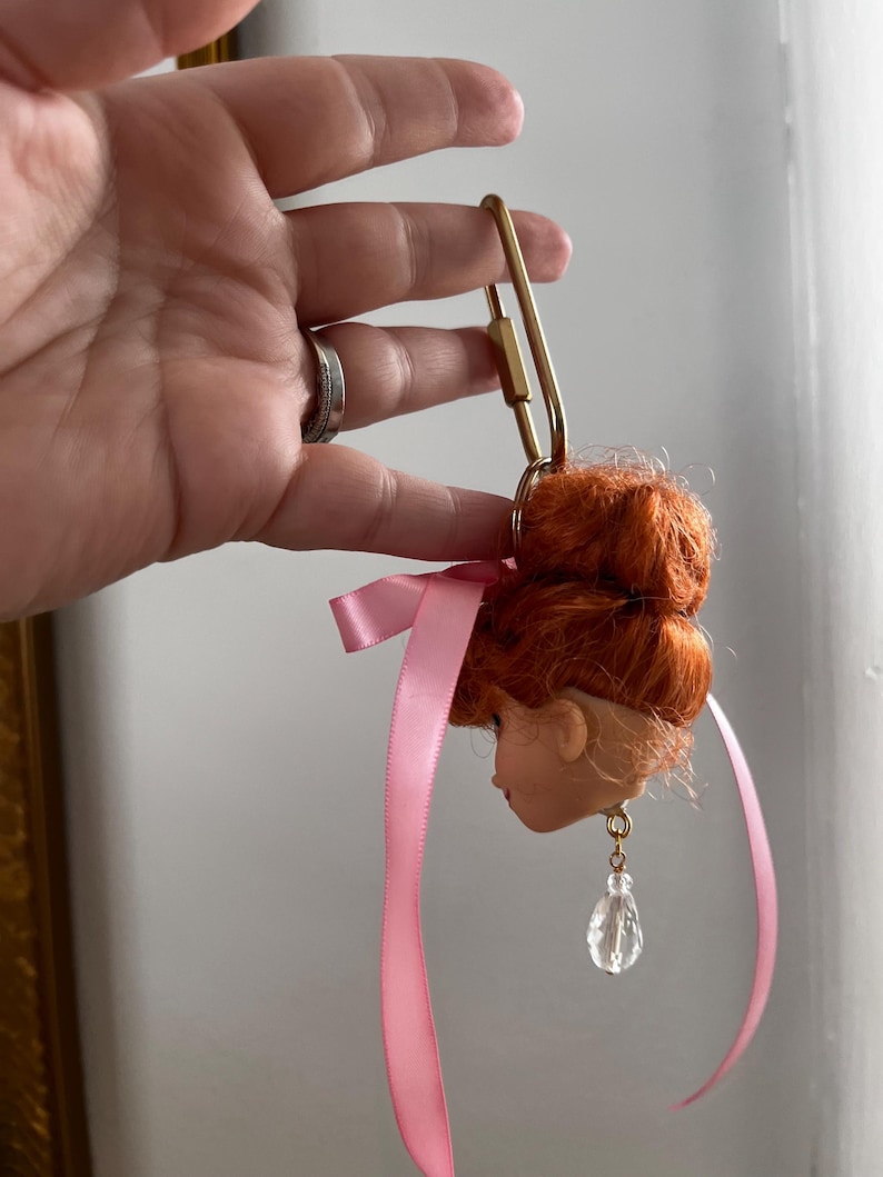 Puede incluir: Una cabeza de mu&ntilde;eca con pelo rojo y una cinta rosa, sujeta a un cierre dorado. La cabeza de la mu&ntilde;eca tiene un pendiente transparente en forma de l&aacute;grima. El cierre est&aacute; sujeto por una mano.