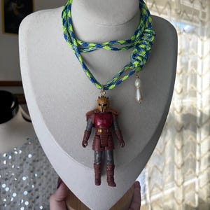 Op de afbeelding: Een unieke ketting met een gevlochten neon groen en blauw koord. De hanger is een klein actiefiguur van een personage in rode en bruine bepantsering. Een witte, druppelvormige kraal hangt aan het koord.