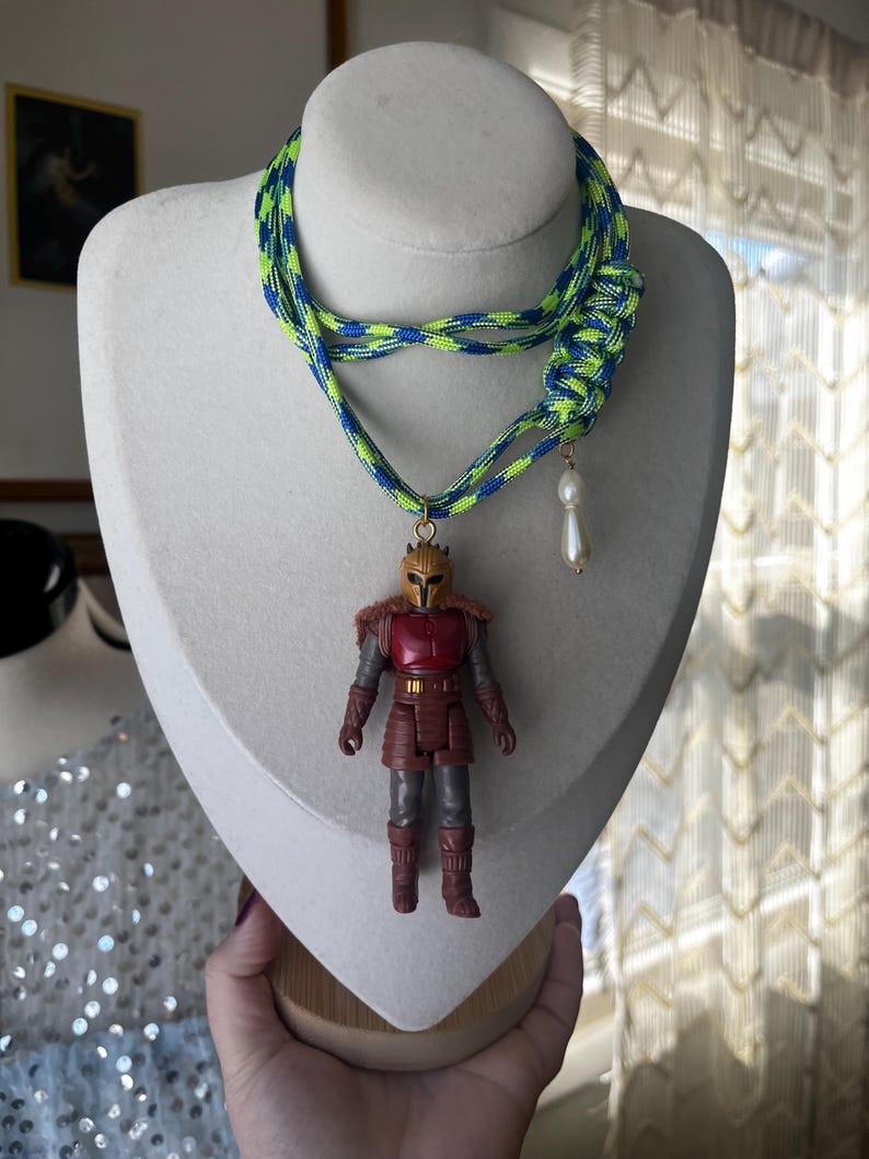 Op de afbeelding: Een unieke ketting met een gevlochten neon groen en blauw koord. De hanger is een klein actiefiguur van een personage in rode en bruine bepantsering. Een witte, druppelvormige kraal hangt aan het koord.