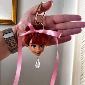 Puede incluir: Un llavero de cabeza de mu&ntilde;eca con cabello rojo, ojos azules y una cinta rosa. El llavero tiene un cierre dorado y un colgante de cristal transparente. La cabeza de la mu&ntilde;eca es un personaje de una pel&iacute;cula de animaci&oacute;n popular.