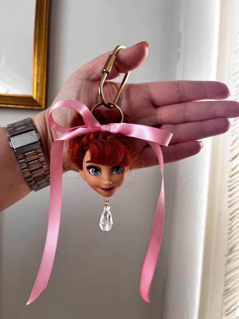 Puede incluir: Un llavero de cabeza de mu&ntilde;eca con cabello rojo, ojos azules y una cinta rosa. El llavero tiene un cierre dorado y un colgante de cristal transparente. La cabeza de la mu&ntilde;eca es un personaje de una pel&iacute;cula de animaci&oacute;n popular.