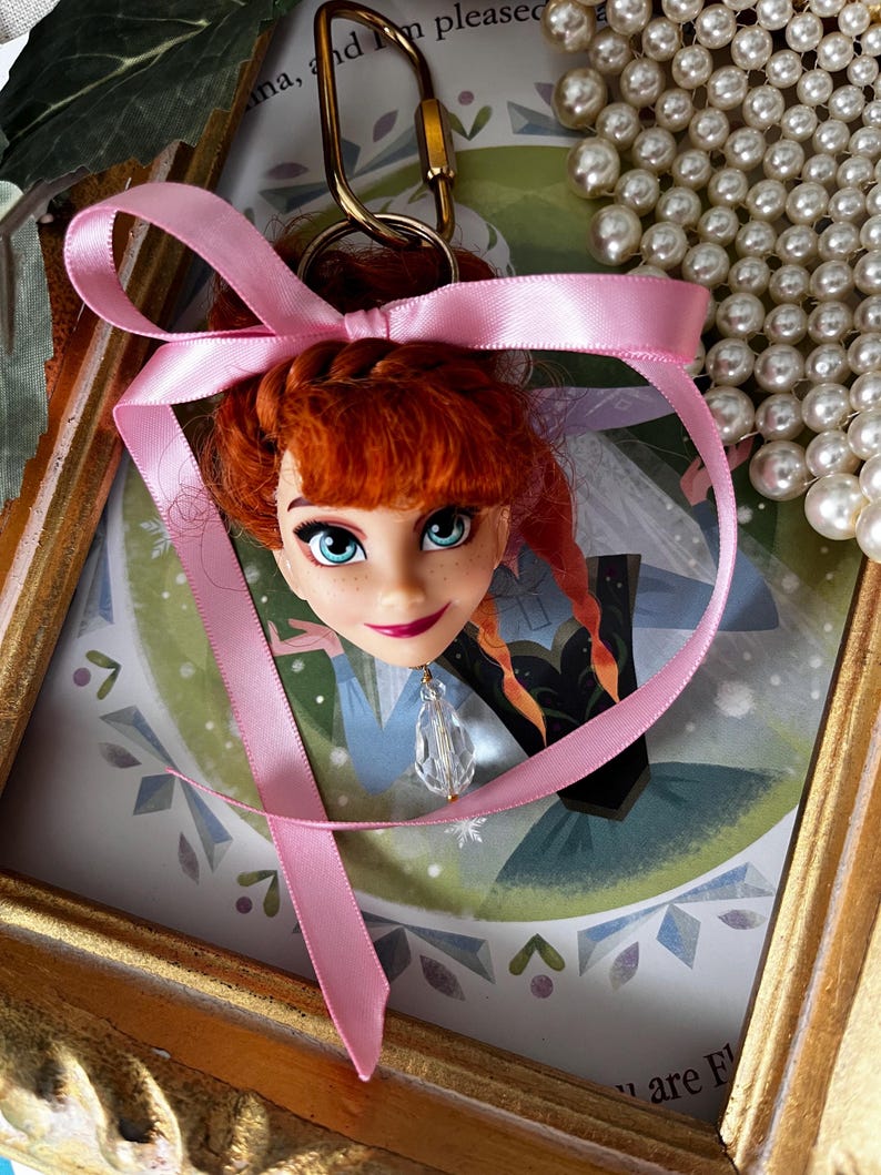 Puede incluir: Un llavero con cabeza de mu&ntilde;eca que presenta un personaje con cabello rojo, ojos azules y una cinta rosa. El llavero est&aacute; sujeto a un cierre dorado. La cabeza de la mu&ntilde;eca est&aacute; colocada contra una imagen enmarcada con un borde dorado.