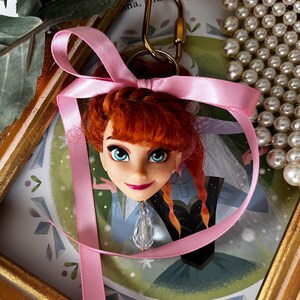 Puede incluir: Un llavero con cabeza de mu&ntilde;eca que presenta un personaje con cabello rojo, ojos azules y una cinta rosa. El llavero tiene un cierre dorado y un colgante de cristal transparente. El fondo incluye una ilustraci&oacute;n enmarcada y un collar de perlas.