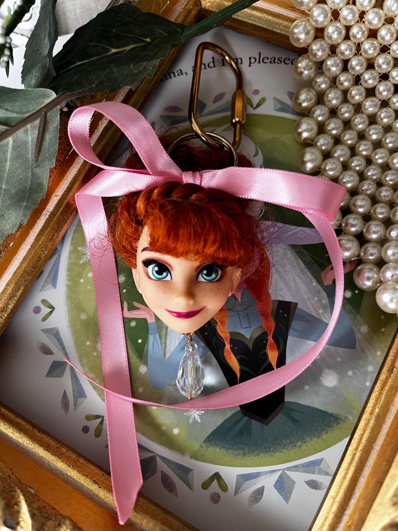 Puede incluir: Un llavero con cabeza de mu&ntilde;eca que presenta un personaje con cabello rojo, ojos azules y una cinta rosa. El llavero tiene un cierre dorado y un colgante de cristal transparente. El fondo incluye una ilustraci&oacute;n enmarcada y un collar de perlas.