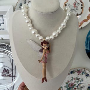 May include: A white pearl necklace with a fairy figurine pendant. The fairy is wearing a pink dress and has brown hair and wings.