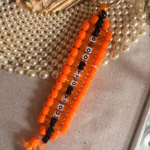 May include: A keychain crafted from vibrant orange and black plastic pony beads arranged in a rectangular pattern. A central column of white letter beads spells out "SPOOKY BOO" vertically. The piece is attached to a circular gold-toned metal ring.
