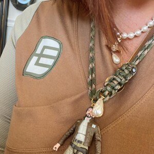 Op de afbeelding: Een bruine en tan ketting met een miniatuur actiefiguur hanger. De ketting heeft een camouflage koord en een parel accent. Een tweede ketting met parels is ook zichtbaar.