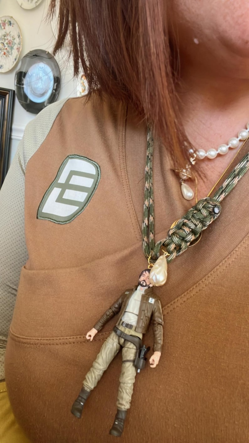 Op de afbeelding: Een bruine en tan ketting met een miniatuur actiefiguur hanger. De ketting heeft een camouflage koord en een parel accent. Een tweede ketting met parels is ook zichtbaar.