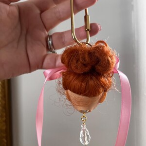Puede incluir: Una cabeza de mu&ntilde;eca con pelo rojo, un cierre dorado y una cinta rosa. Un cristal transparente en forma de l&aacute;grima cuelga de la cabeza de la mu&ntilde;eca. Un art&iacute;culo &uacute;nico y artesanal.