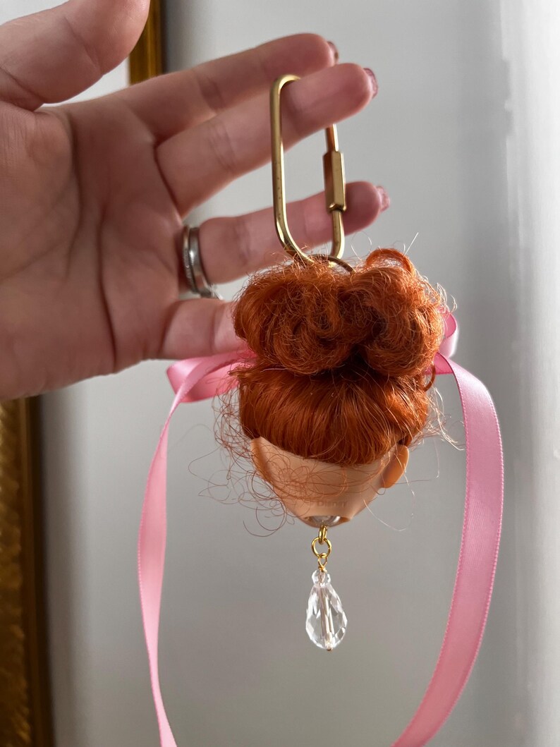 Puede incluir: Una cabeza de mu&ntilde;eca con pelo rojo, un cierre dorado y una cinta rosa. Un cristal transparente en forma de l&aacute;grima cuelga de la cabeza de la mu&ntilde;eca. Un art&iacute;culo &uacute;nico y artesanal.