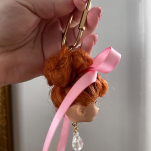Puede incluir: Un llavero con cabeza de mu&ntilde;eca con pelo rojo recogido en un mo&ntilde;o, una cinta rosa y un cierre dorado. La mu&ntilde;eca tiene un pendiente de l&aacute;grima de cristal transparente. Un accesorio &uacute;nico.