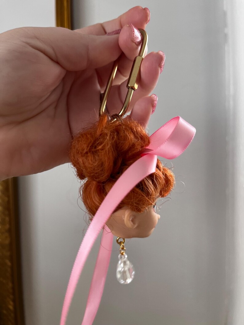 Puede incluir: Un llavero con cabeza de mu&ntilde;eca con pelo rojo recogido en un mo&ntilde;o, una cinta rosa y un cierre dorado. La mu&ntilde;eca tiene un pendiente de l&aacute;grima de cristal transparente. Un accesorio &uacute;nico.