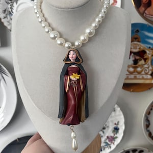 May include: A pearl necklace with a pendant featuring a figure in a hooded cloak holding a yellow flower. The figure is wearing a burgundy dress and a black cloak. A single pearl dangles from the bottom of the pendant.