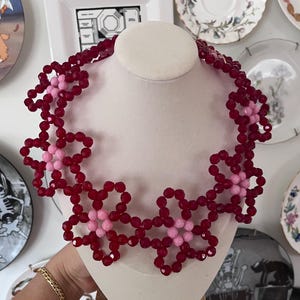 May include: A red and pink beaded necklace with a flower design. The necklace is made of small, faceted beads and is arranged in a pattern of overlapping flowers.