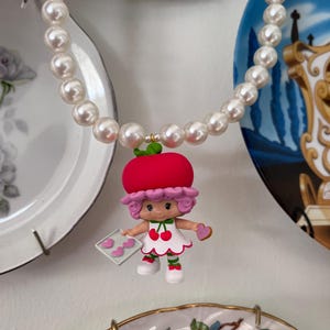May include: A white pearl necklace with a red and white strawberry shortcake doll charm. The doll is holding a pink baking sheet with pink heart-shaped cookies.