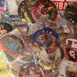 May include: A collection of circular Disney-themed pinback buttons, each individually wrapped in clear plastic and accented with small white pearls. The designs feature various characters and attractions, including Mickey Mouse, Donald Duck, and Haunted Mansion.