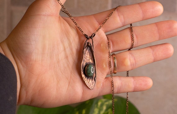 A real cicada wing — iridescent, impossibly delicate — preserved in electroformed copper with an emerald set inside it; genuinely a 'how did they even make this?' piece.