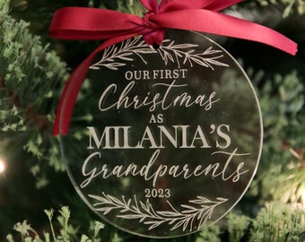 May include: A circular clear acrylic Christmas ornament featuring a red satin ribbon hanger. The text reads: OUR FIRST Christmas AS MILANIA'S Grandparents 2023. The design includes etched botanical branch illustrations at the top and bottom.