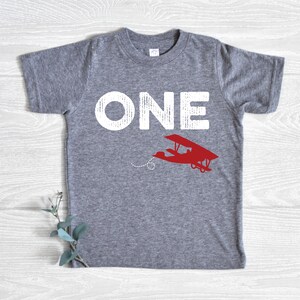 May include: A gray t-shirt with the word "ONE" in distressed white lettering and a red airplane graphic. The airplane is flying with a dotted line behind it. The shirt is a children's size.