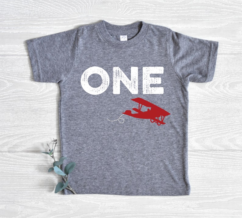 May include: A gray t-shirt with the word "ONE" in distressed white lettering and a red airplane graphic. The airplane is flying with a dotted line behind it. The shirt is a children's size.