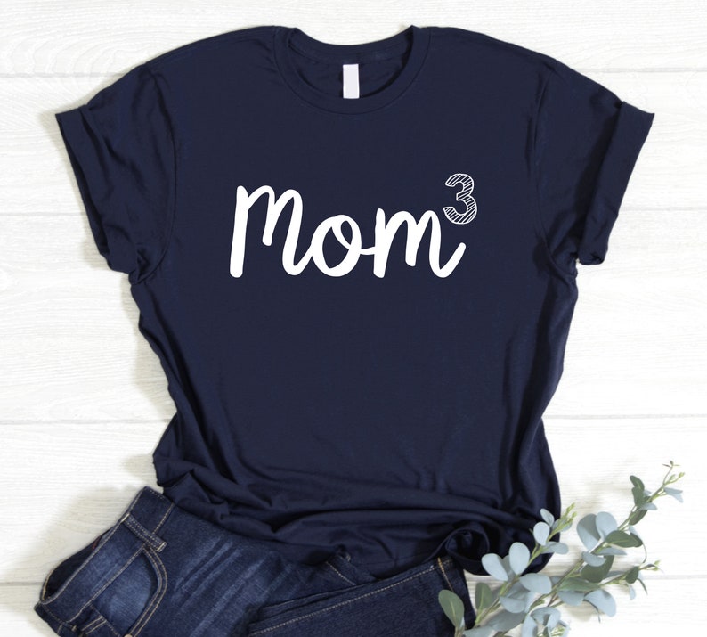May include: A navy blue t-shirt featuring the word "Mom" in white script, with a white, outlined "3" as an exponent. The shirt is folded, with blue jeans visible below. A sprig of greenery is in the lower right corner.