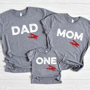 May include: Three gray t-shirts with white text and a red airplane graphic. The shirts read "DAD", "MOM", and "ONE". The shirts are likely for a family, with the "ONE" shirt being for a child.