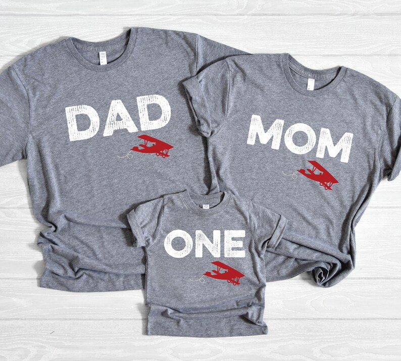 May include: Three gray t-shirts with white text and a red airplane graphic. The shirts read "DAD", "MOM", and "ONE". The shirts are likely for a family, with the "ONE" shirt being for a child.