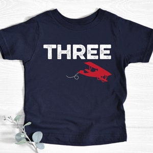 May include: Navy blue toddler t-shirt with the word "THREE" in white block letters. A red airplane graphic is below the word "THREE". The airplane has a dotted line trailing behind it. The shirt is made of soft material.
