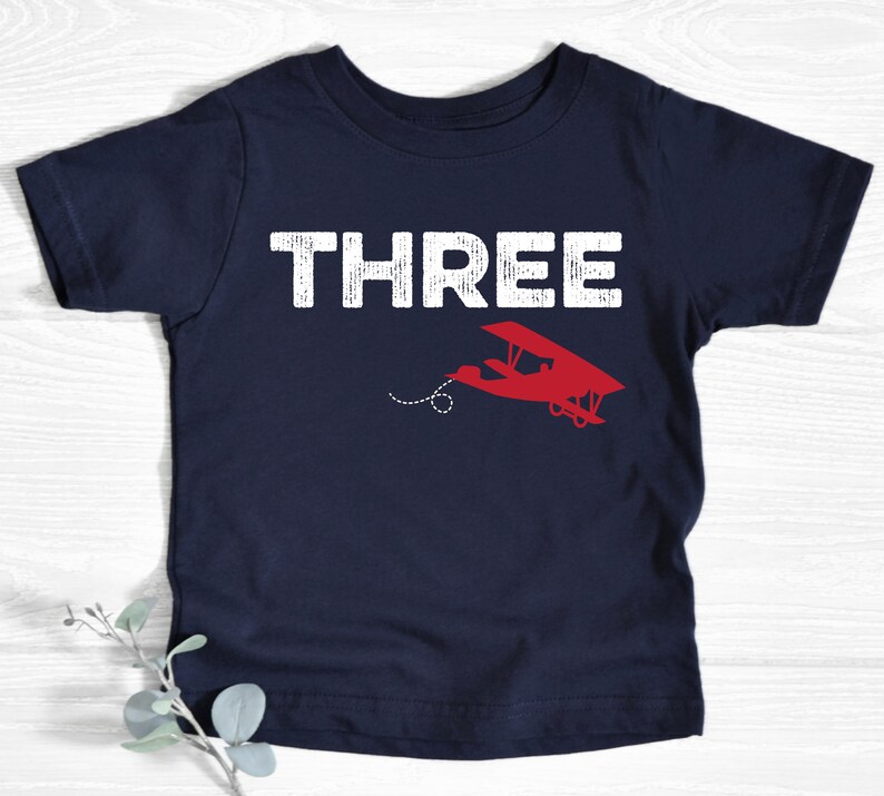May include: Navy blue toddler t-shirt with the word "THREE" in white block letters. A red airplane graphic is below the word "THREE". The airplane has a dotted line trailing behind it. The shirt is made of soft material.