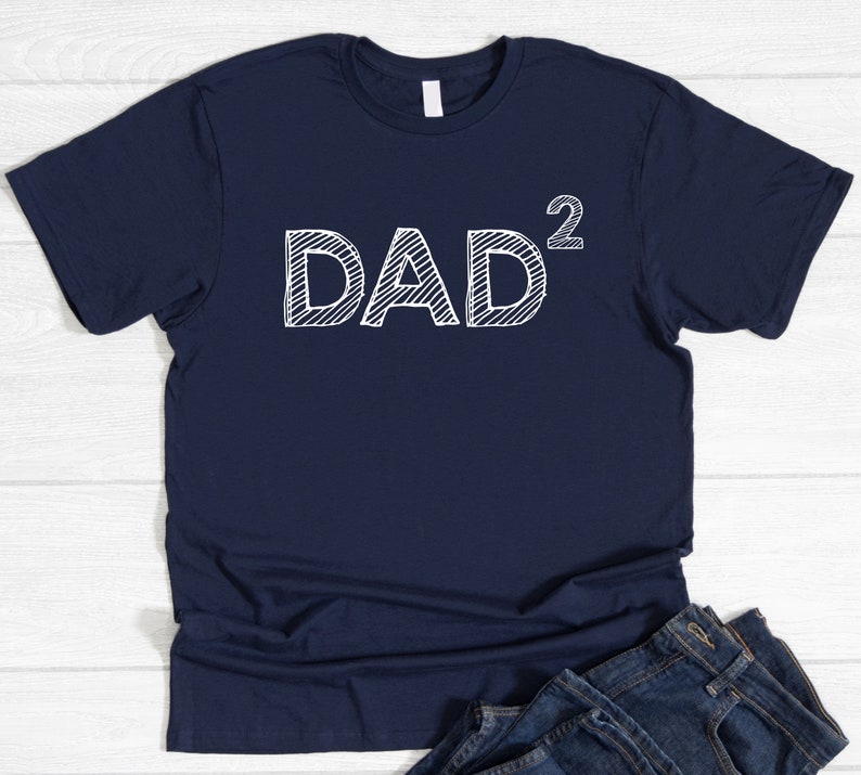 May include: A navy blue t-shirt featuring the white text "DAD&sup2;" in a hand-drawn style. The shirt is folded, with blue jeans visible at the bottom. The design is centered on the front of the tee.