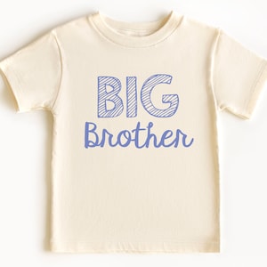 May include: Cream-colored toddler t-shirt with the words "BIG Brother" printed in blue. The word "BIG" is in a block letter style with diagonal lines, and "Brother" is in a cursive font.