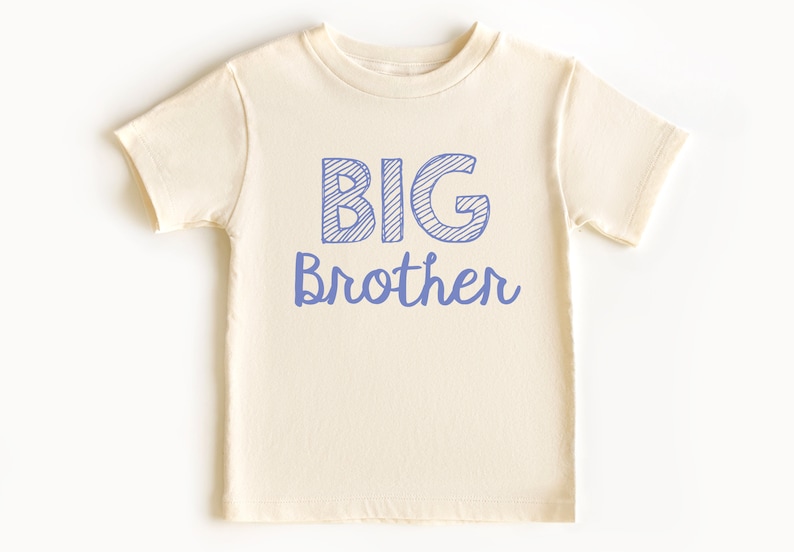May include: Cream-colored toddler t-shirt with the words "BIG Brother" printed in blue. The word "BIG" is in a block letter style with diagonal lines, and "Brother" is in a cursive font.