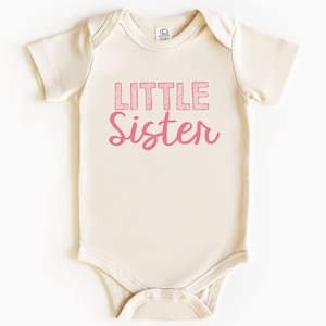 May include: A light beige baby onesie with the words "Little Sister" printed in pink. The word "Little" is in a block letter style with a crosshatch design, and "Sister" is in a cursive font.