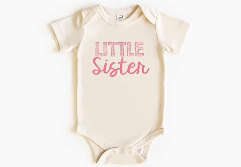 May include: A light beige baby onesie with the words "Little Sister" printed in pink. The word "Little" is in a block letter style with a crosshatch design, and "Sister" is in a cursive font.