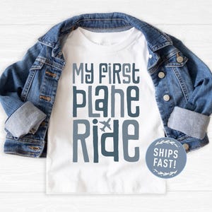 May include: White t-shirt with blue text that reads "My First Plane Ride" with a small airplane graphic. The shirt is laid on a wooden surface with a blue denim jacket folded on top.