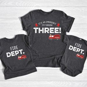 May include: Three matching shirts for a family. The largest shirt is dark gray with white text that reads "It's an emergency I'm turning THREE!" with a red fire truck graphic. The medium shirt is dark gray with white text that reads "FIRE DEPT." with a red fire truck graphic. The smallest shirt is a dark gray onesie with white text that reads "FIRE DEPT." with a red fire truck graphic.